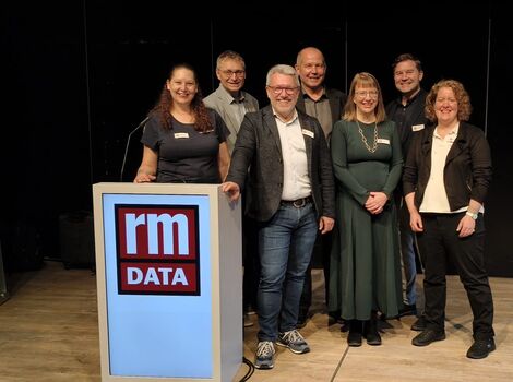 Gruppe von sieben Personen posiert hinter einem Podest mit dem rm DATA-Logo bei einer Firmenveranstaltung; alle tragen geschäftliche Kleidung und lächeln in die Kamera, während im Hintergrund Banner und eine dunkle Bühne zu sehen sind.