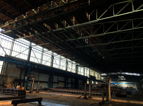 Große Industriehalle mit offener Dachkonstruktion und Stahlträgern. Links erstrecken sich Fensterreihen, durch die Tageslicht hinein fällt. Auf dem Hallenboden liegen gestapelte Metallbahnen und Bauteile; im Hintergrund sind weitere Strukturen und Gerüste sichtbar.