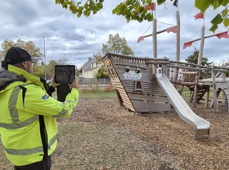 Eine Person in fluoreszierend gelber Warnjacke filmt oder fotografiert ein hölzernes Kletter- und Spielgerät mit Rutsche auf einem Spielplatz; im Hintergrund stehen Bäume, der Himmel ist wolkig.