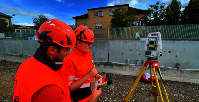 Zwei Bauarbeiter in leuchtend orange Warnkleidung und roten Helmen arbeiten auf einer Baustelle und bedienen ein Messgerät auf einem Stativ; im Hintergrund sind Gebäude und eine Betonmauer sichtbar.