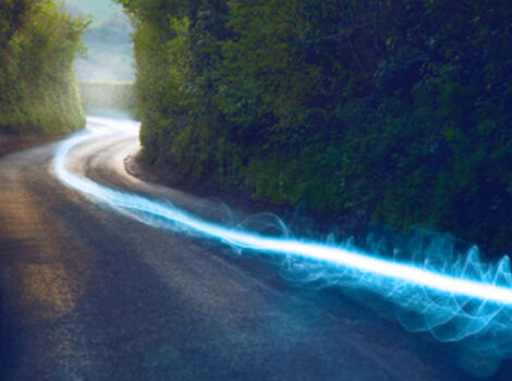 Eine kurvige Straße windet sich durch einen dicht bewaldeten Wald; Bäume säumen beide Seiten, während eine leuchtend blaue Lichtspur sich entlang der Fahrbahn zieht.