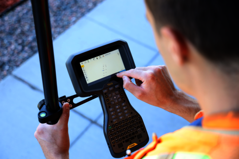Eine Person in einer leuchtenden Sicherheitsweste bedient ein tragbares Datenterminal mit Tastatur und Touchscreen; eine Hand hält das Gerät, die andere tippt auf dem Bildschirm.