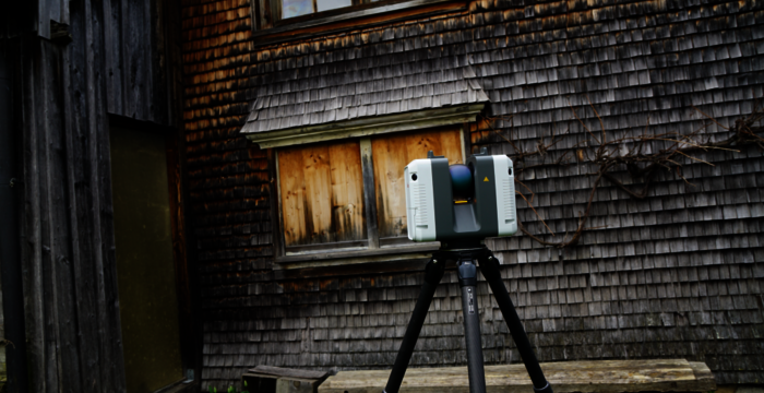 Ein Vermessungsgerät (Totalstation) auf einem Stativ steht vor einem alten Holzhäuschen.