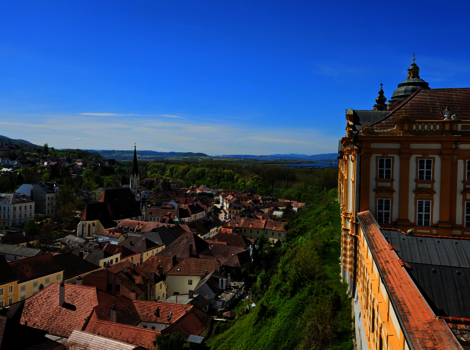 Ausblick über eine malerische Stadt mit roten Ziegeldächern, Kirchtürmen und engen Gassen. Rechts im Vordergrund ein prachtvolles Barockgebäude, links grüne Hügel und ein weiter blauer Himmel.