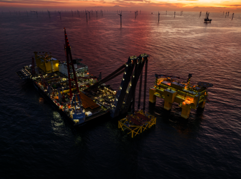 Offshore-Bohrplattform im Meer bei Sonnenuntergang. Gelbe Strukturteile und ein großer Kran ragen aus dem Wasser, Beleuchtung leuchtet. Im Hintergrund sind weitere Ölplattformen und Pfähle zu sehen. Ruhiges Wasser spiegelt den warmen, orangefarbenen Himmel.