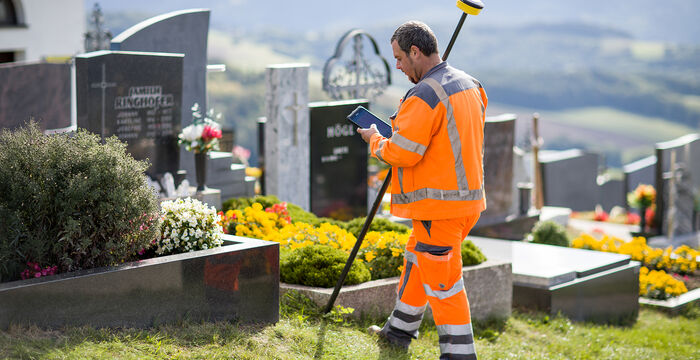 Ein Friedhofsarbeiter in leuchtend orangefarbener Warnkleidung pflegt das Gras um Grabsteine. Er nutzt ein langstieliges Gartenwerkzeug; Blumen schmücken die Gräber, im Hintergrund erstrecken sich Felder und sanfte Hügel.