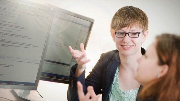Software-Entwicklung Zwei Personen arbeiten an einem Computer. Eine Frau mit Brille erklärt etwas und gestikuliert, während die andere zuhört. Auf dem Monitor sind Programmierzeilen sichtbar.