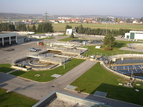 Luftaufnahme eines Klärwerks: mehrere runde Becken mit Rohrleitungen, angrenzende Wege und Grünflächen; im Hintergrund eine kleine Stadt mit Häusern.